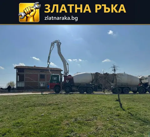 Разгънатата стрела на бетонпомпа достига до тухлена сграда до два бетоновоза на открит терен