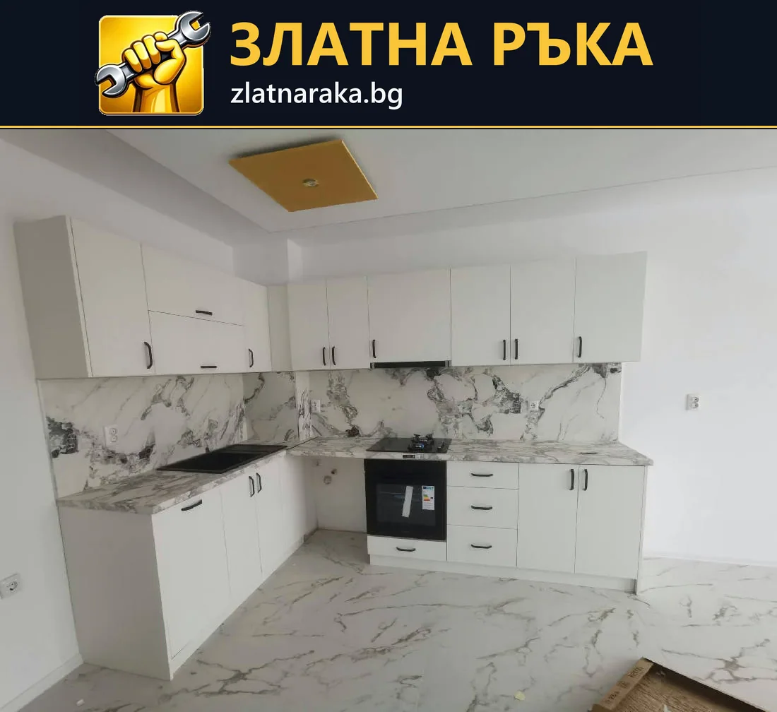 Бяла ъглова кухня с горни шкафове, вградена фурна и светъл гръб с мраморен рисунък