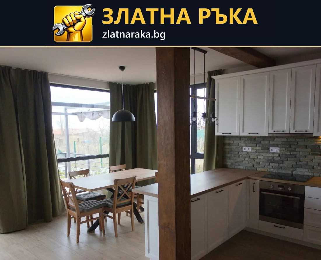 Ъглова бяла кухня с дървесен плот и тухлен гръб до маса край прозорец.