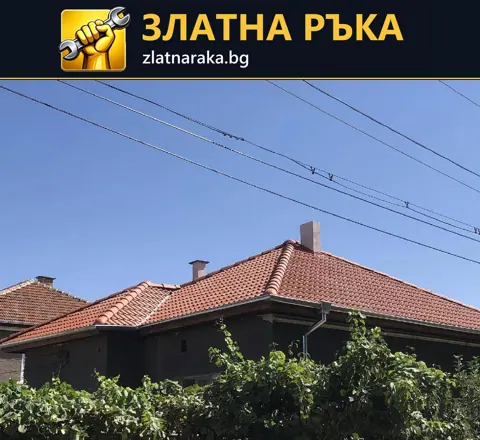 Къща с червен керемиден покрив, изразени билни линии и тъмна дървена фасада зад дворна зеленина