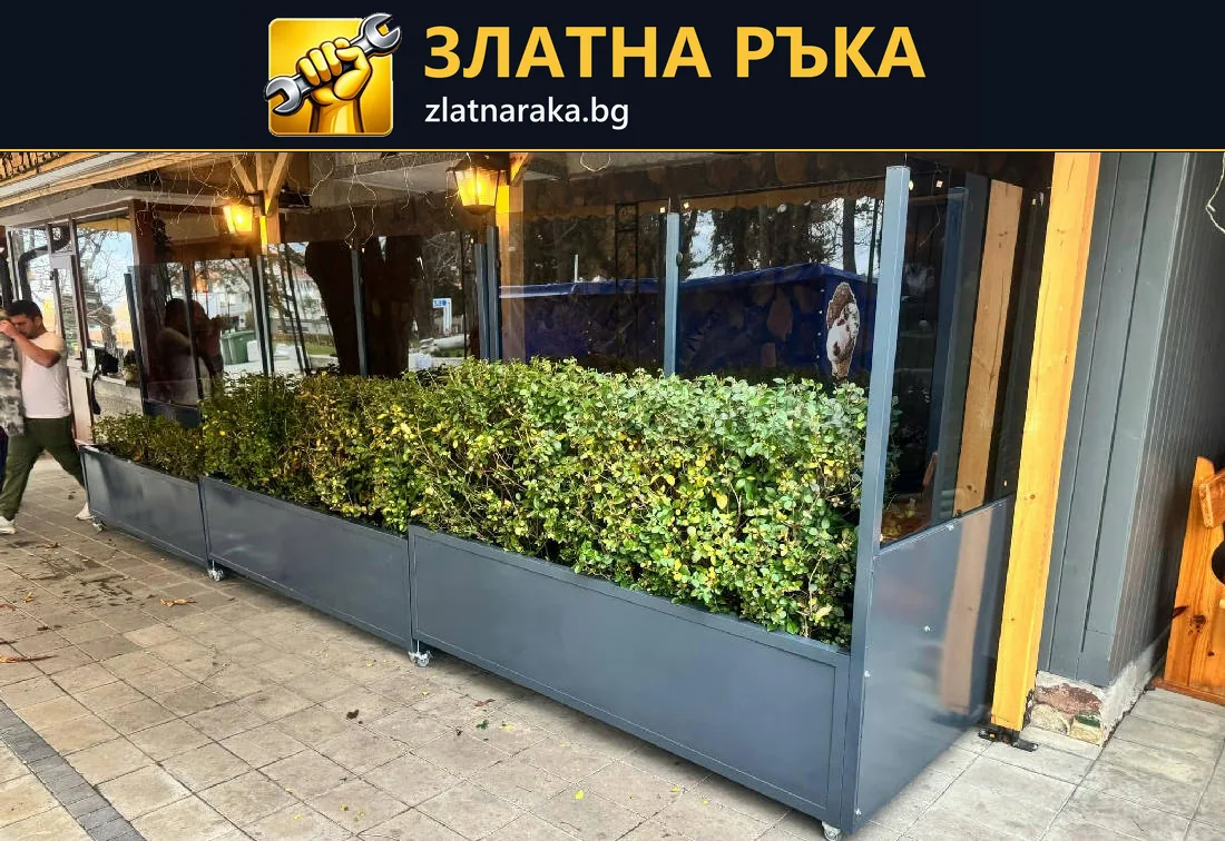 Редица правоъгълни тъмносиви метални цветарници с гъста зеленина върху настилка пред заведение