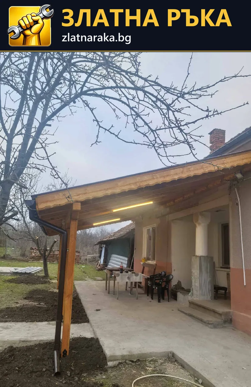 Дълъг дървен навес с видими колони покрива бетонна площадка и кът за сядане до къща