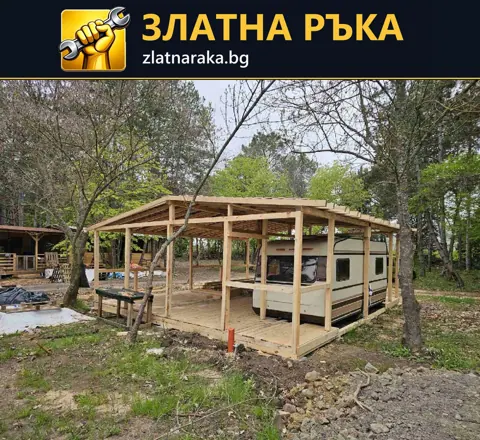 Дървени колони и покривни греди оформят навес около каравана сред дървета.