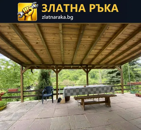 Покрита веранда с дървени колони и греди, квадратни настилъчни плочи и маса с пейки до зелен двор