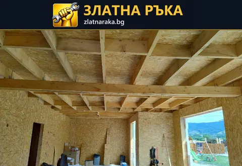 Дървени греди по тавана и стенни плоскости в строящо се помещение с голям прозоречен отвор.