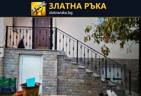 Външно стълбище към тъмна входна врата с черни метални балюстри и сива облицовка по високия цокъл