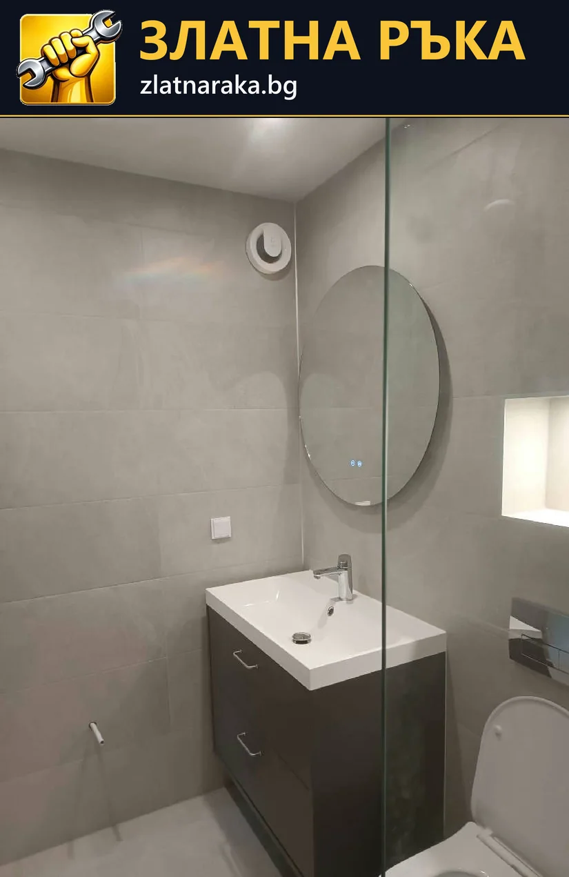 Прозрачен стъклен параван пресича изгледа към тъмен шкаф с мивка и кръгло огледало върху сива облицовка