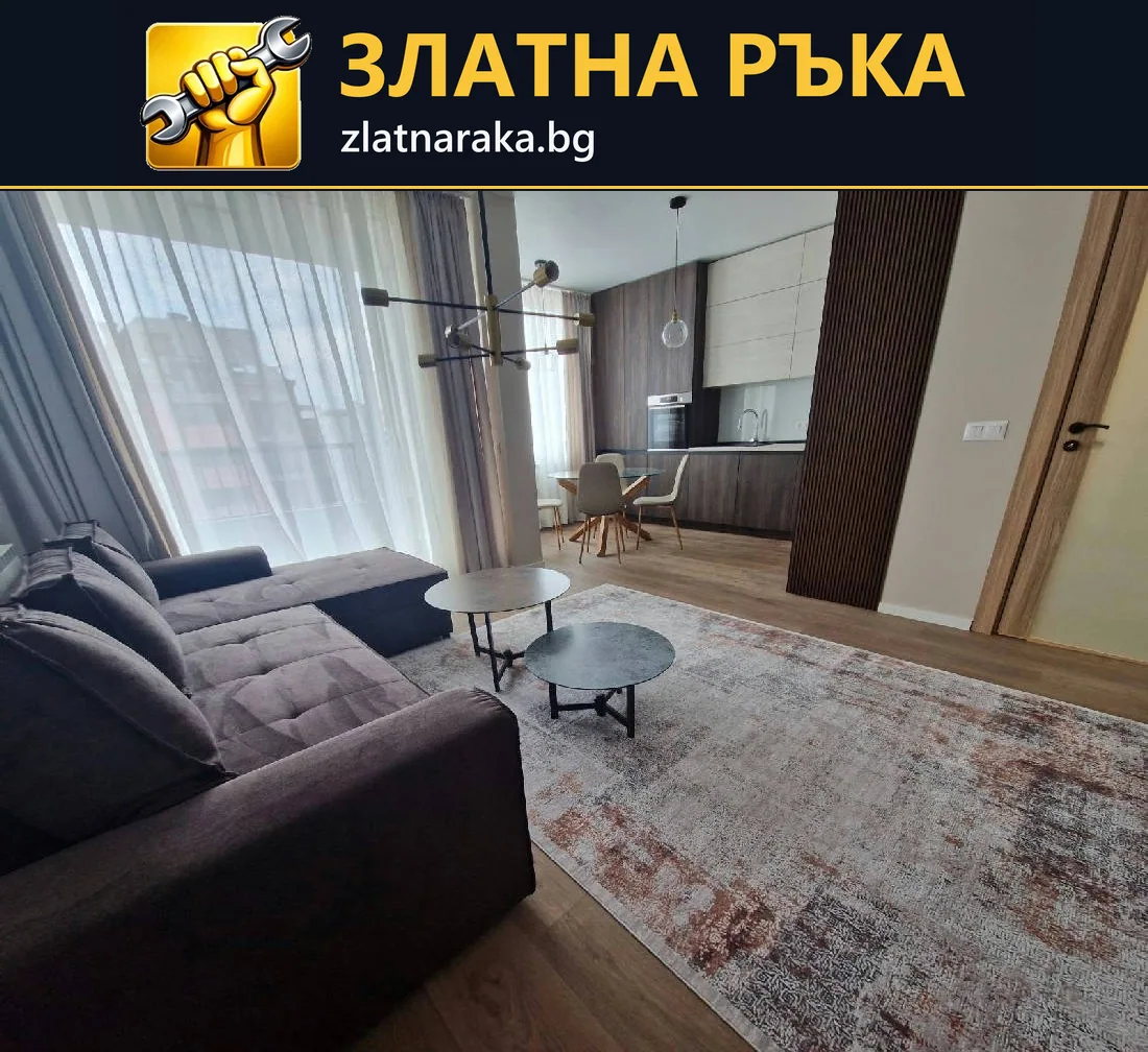 Светла дневна с тъмен ъглов диван, голям килим и кухненска зона зад ниски холни маси