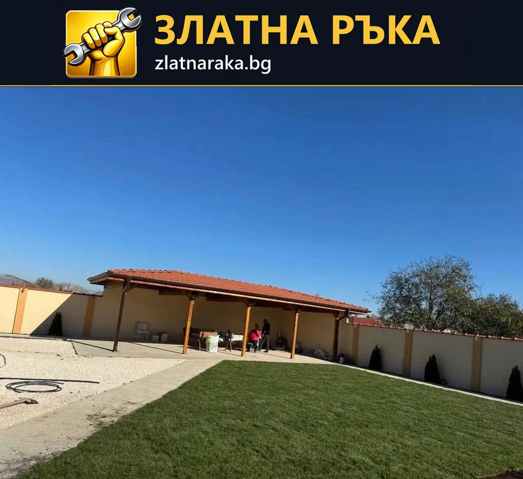 Зелена правоъгълна морава е отделена от светла дворна настилка пред дълъг навес с червен покрив