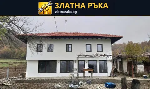 Бяла двуетажна къща с червен покрив, тъмни прозоречни рамки и незавършено пространство пред фасадата
