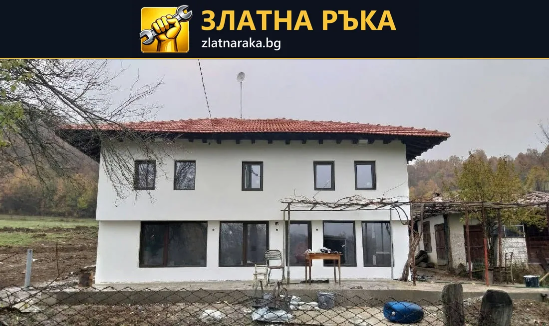 Бяла двуетажна къща с червен покрив, тъмни прозоречни рамки и незавършено пространство пред фасадата