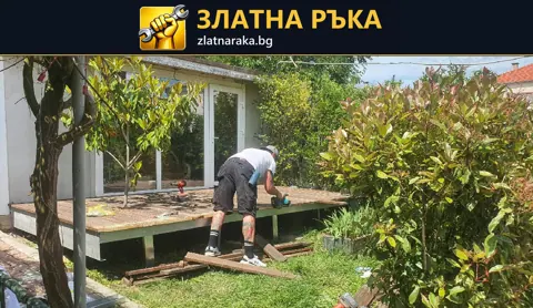 Работник оформя дървена площадка с открита челна конструкция сред дървета и храсти.