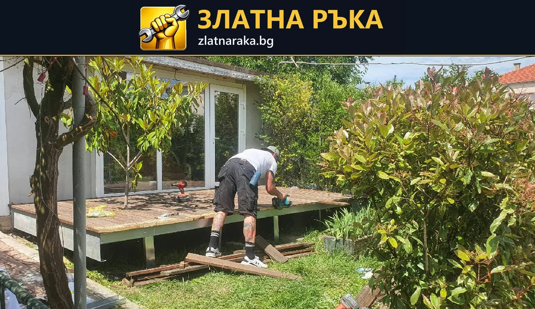 Работник оформя дървена площадка с открита челна конструкция сред дървета и храсти.