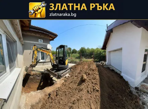 Малък багер до открит изкоп и натрупана почва между две светли сгради
