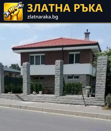 Двуетажна къща с червен покрив стои зад високи светли колони и ниска основа с релефна облицовка