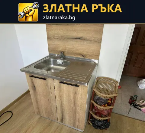 Малка метална мивка с отцедник е поставена върху двукрилен шкаф със светъл дървесен декор