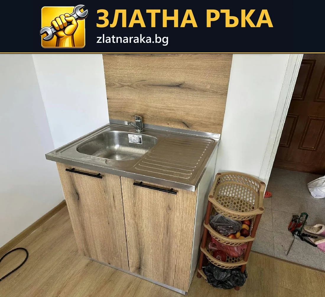 Малка метална мивка с отцедник е поставена върху двукрилен шкаф със светъл дървесен декор