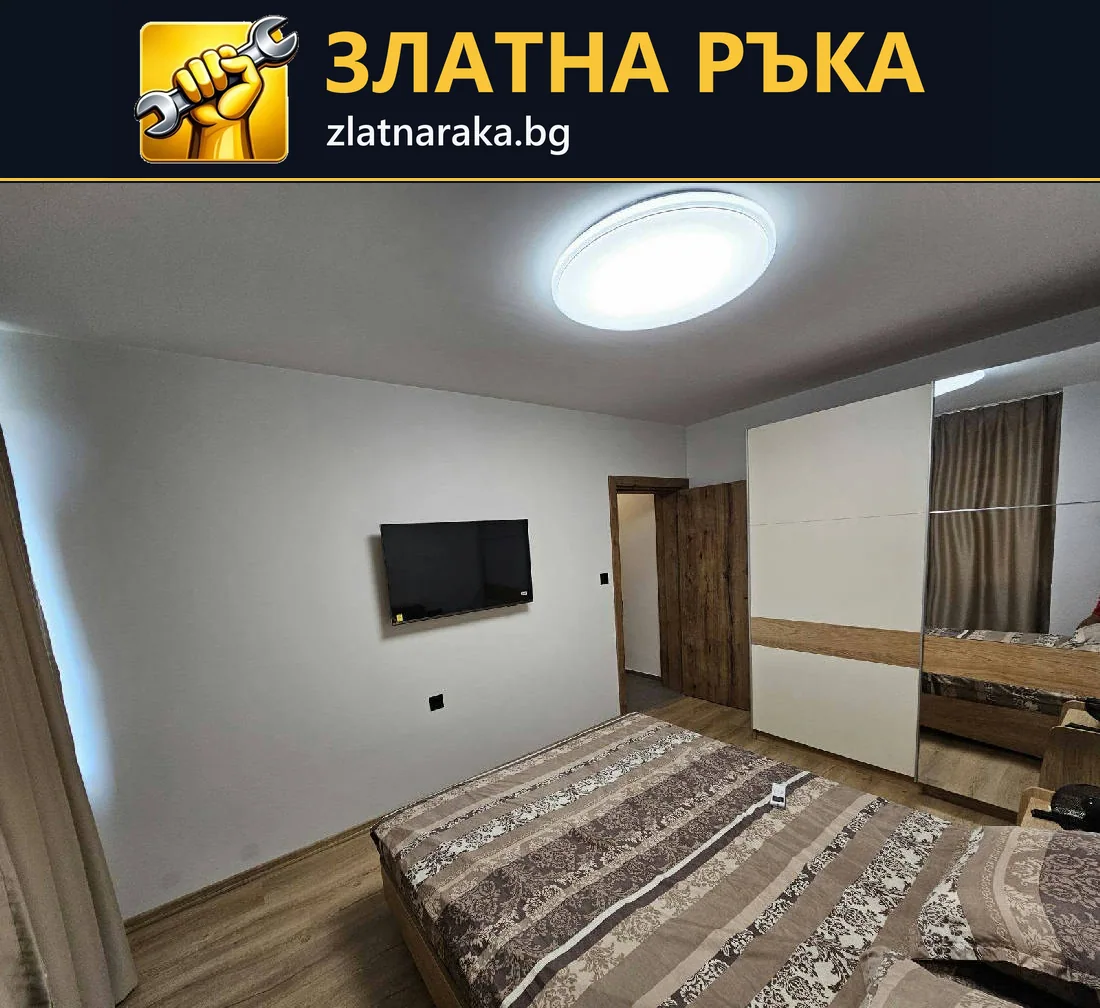 Обзаведена спалня с голям гардероб, стенен телевизор и кръгло таванно осветление.