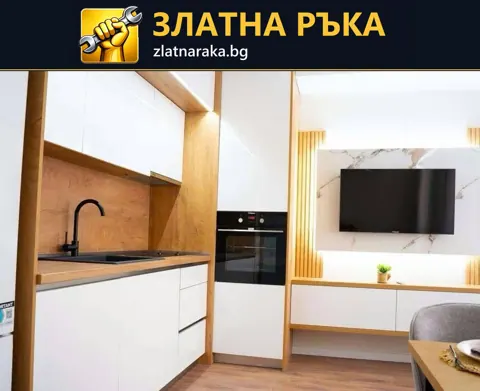 Бяла кухня с вградена фурна и черна мивка продължава към телевизионна стена с дървесни акценти