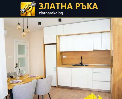 Линейна кухня с бели фронтове, дървесен гръб, черен смесител и хладилник до малка трапезна маса