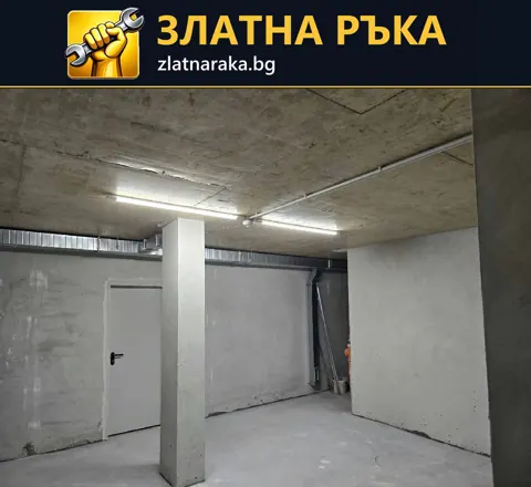 Бетонно помещение с дълги таванни лампи, открити тръбни трасета и квадратни колони.