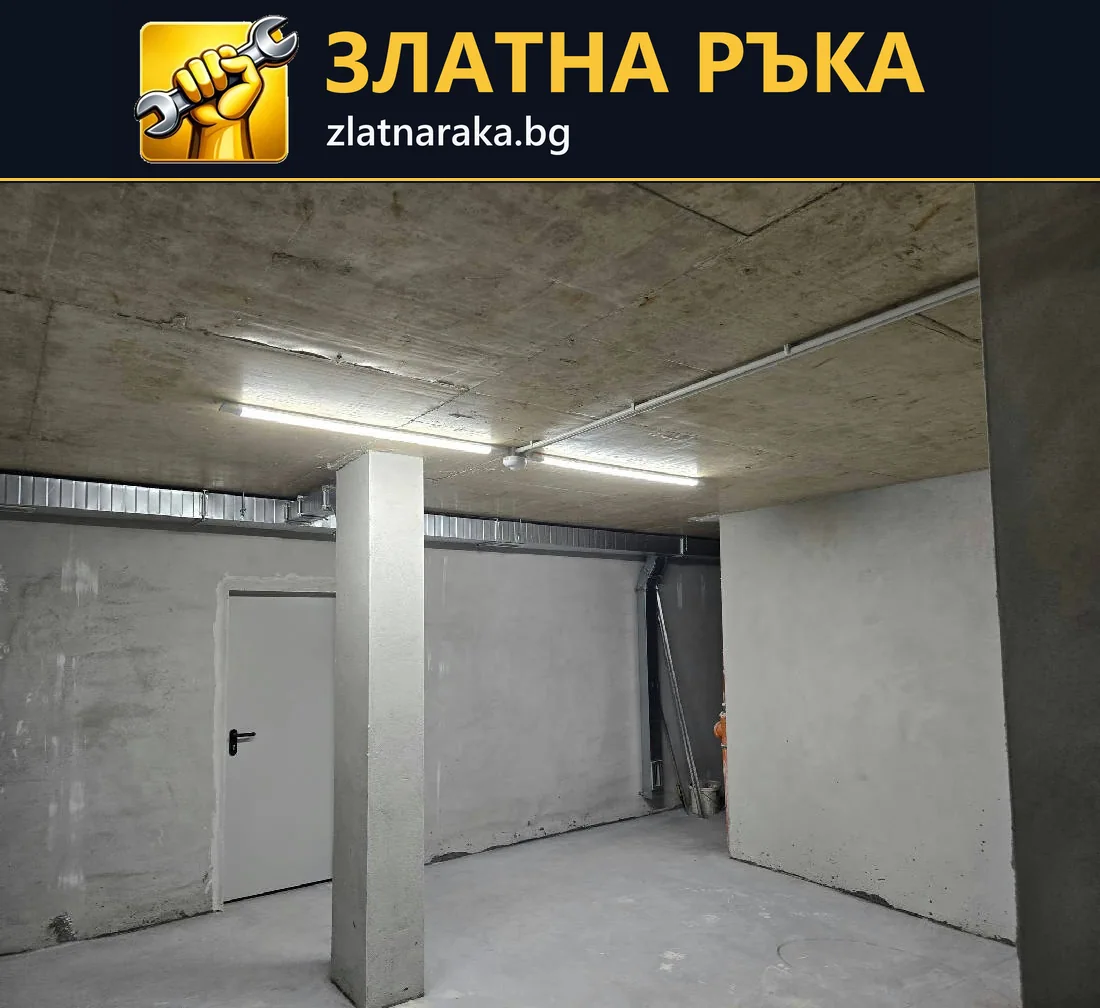 Бетонно помещение с дълги таванни лампи, открити тръбни трасета и квадратни колони.