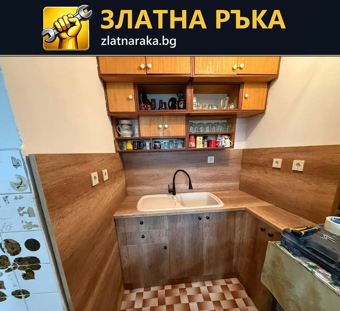Ъглов кухненски плот с бяла мивка, тъмен смесител и дървесни шкафове под открити горни рафтове
