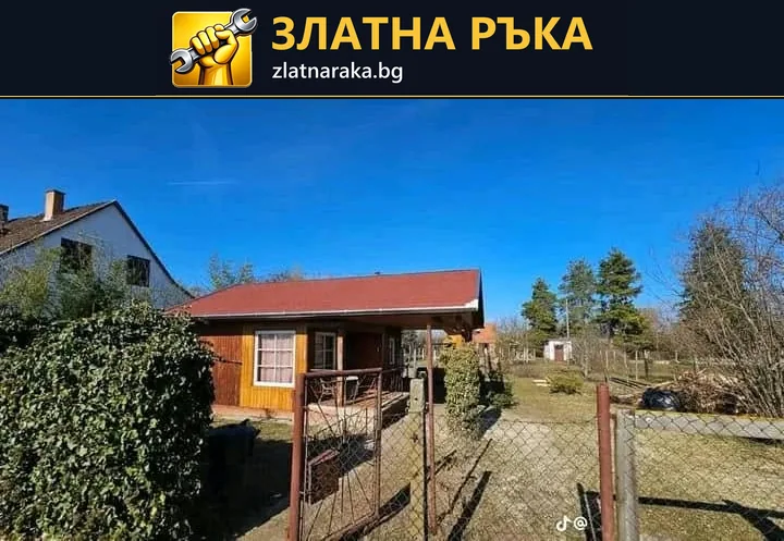 Дървена постройка с червен едноскатен покрив, прозорец и малка покрита веранда стои в ограден двор