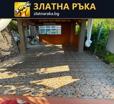 Пъстра настилка с неправилни фигури обикаля ствол на дърво под дървен навес и зелена ограда