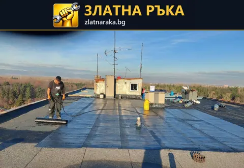 Работник с дълъг инструмент разнася тъмно покритие по плосък покрив около ниски покривни постройки