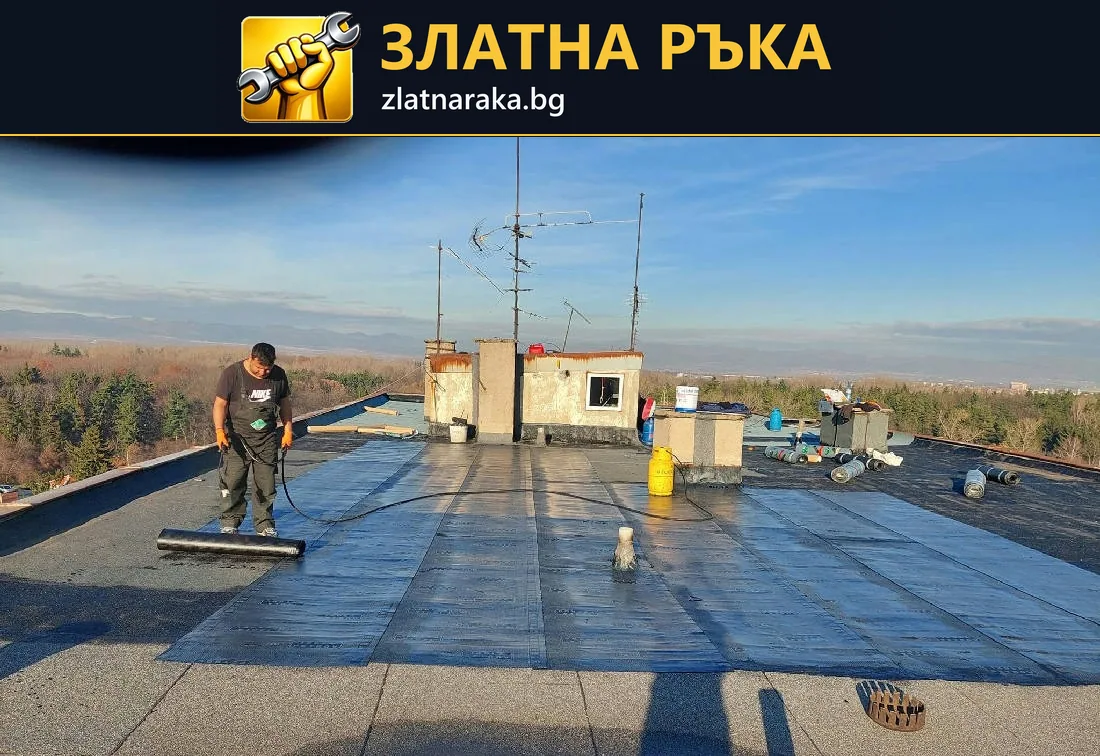 Работник с дълъг инструмент разнася тъмно покритие по плосък покрив около ниски покривни постройки
