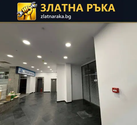 Широк коридор в търговска сграда с бели колони, тъмни подови плочки и редове таванни лунички