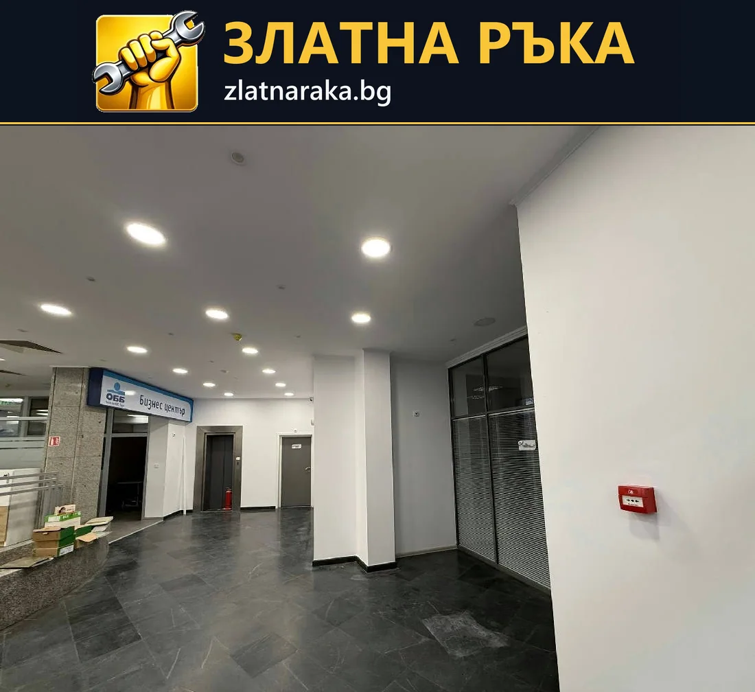 Широк коридор в търговска сграда с бели колони, тъмни подови плочки и редове таванни лунички