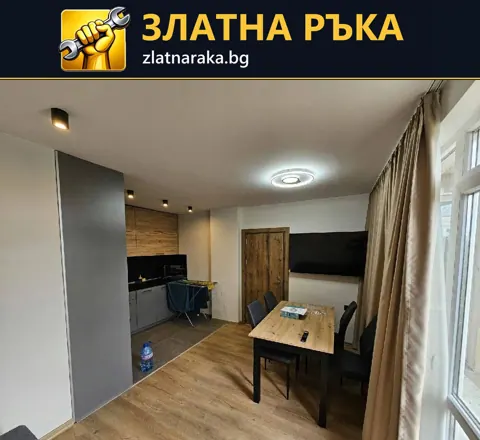 Дневна с дървесен под, трапезна маса и компактна кухня зад сива частична преграда до прозорци
