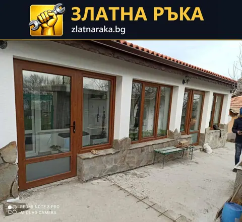 Дълга остъклена фасада с кафява дограма, прозорци и входна врата пред каменен цокъл.
