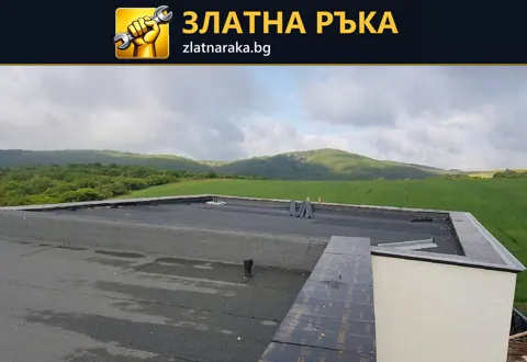 Плосък покрив с тъмна хидроизолация и светли периферни бордове на сграда сред открит терен.