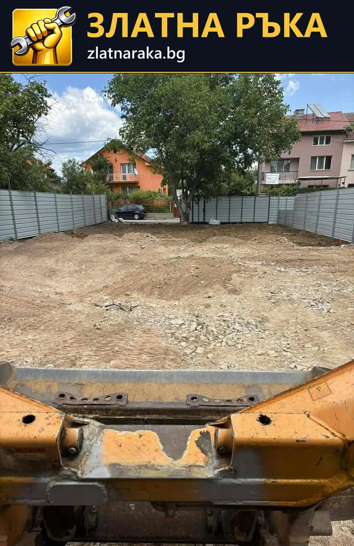 Широка изравнена земна площ зад ограда с част от строителна машина на преден план