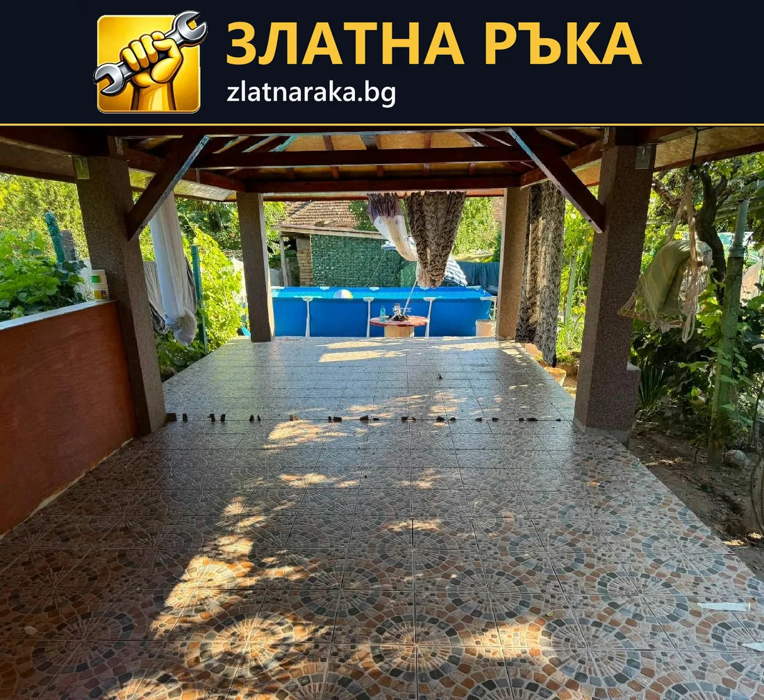 Дълга настилка с разноцветни неправилни парчета под дървен навес води към кът със сини седалки