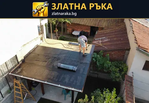 Работник върху плосък дворен навес с частично положено тъмно покритие и руло на покривната площ