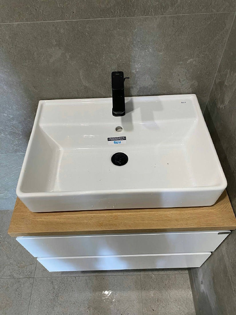Бяла мивка върху дървесен плот и шкаф с бели чекмеджета; видими капки вода по мивката и пода
