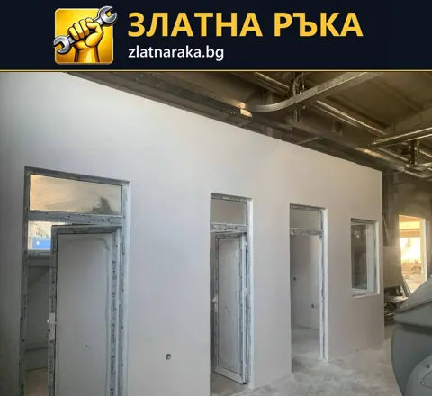 Дълга светла преградна стена с врати и тесни прозорци под видими метални таванни елементи