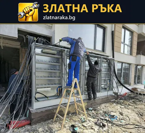 Монтажници работят по големи електрически табла с множество кабели пред фасада на строеж.