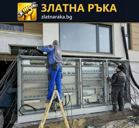 Двама работници свързват кабели към отворено електрическо табло пред сграда