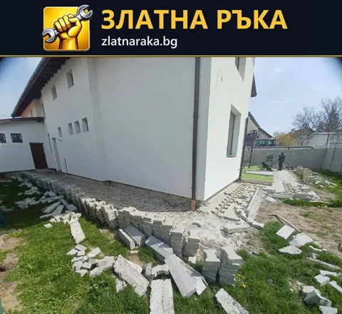 Полагане на сиви павета и бордюри около бяла фасада на къща; материалът е подреден край недовършената пътека.