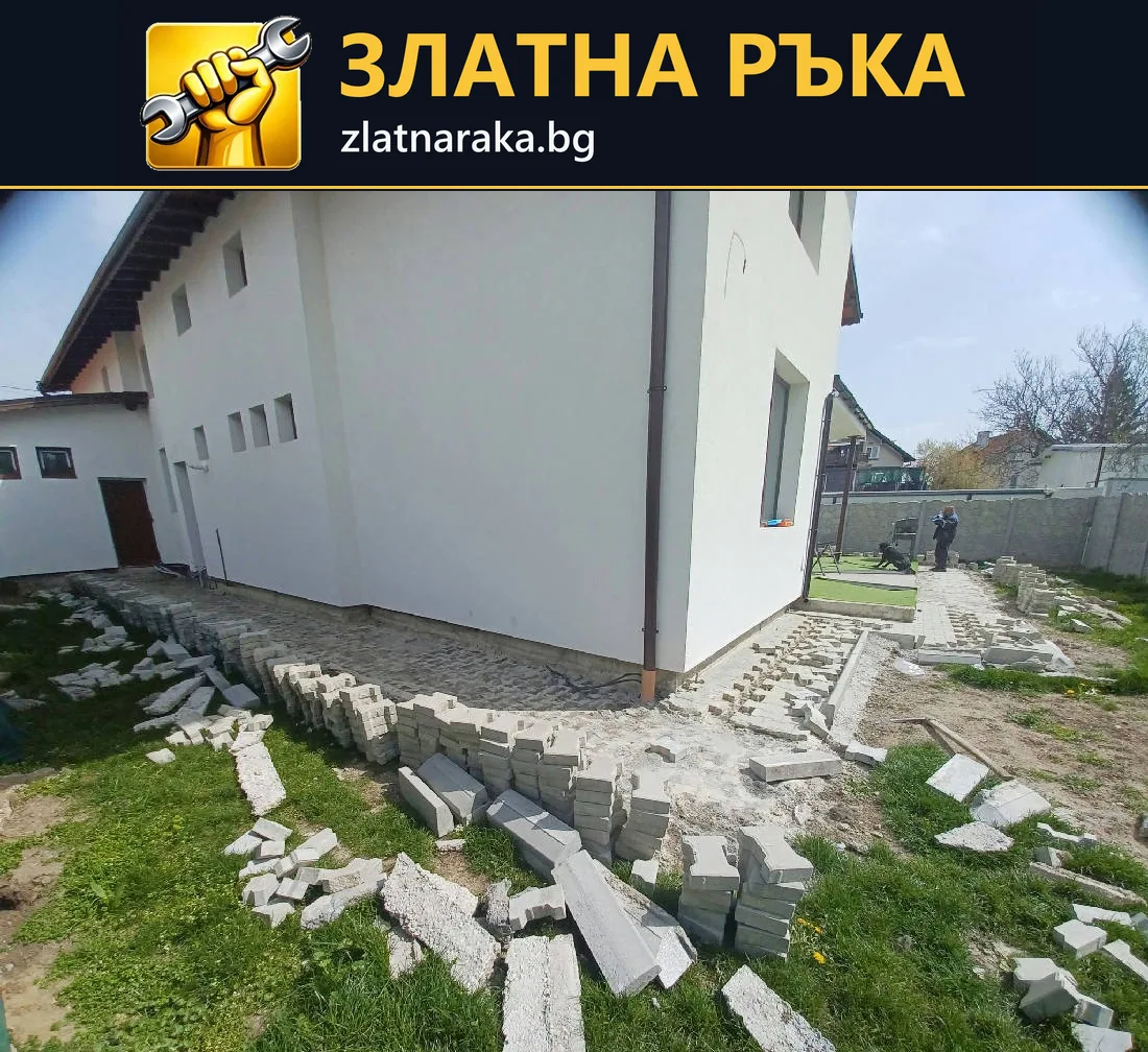 Полагане на сиви павета и бордюри около бяла фасада на къща; материалът е подреден край недовършената пътека.