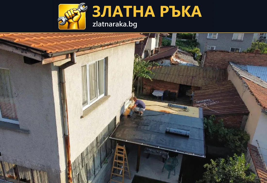 Работник и разстлани рулонни материали са върху нисък покрив, прилепен към светла двуетажна къща