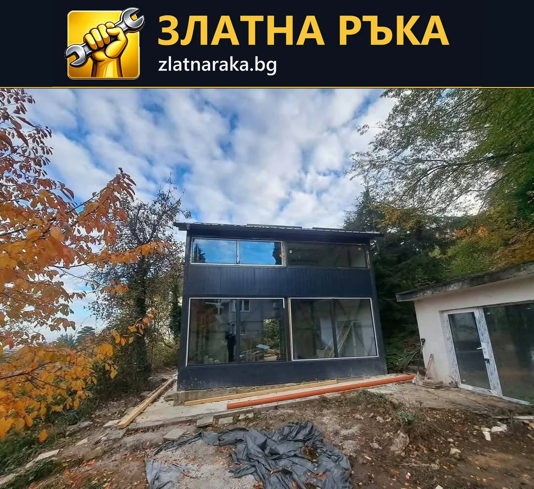 Двуетажна компактна къща с тъмна фасада, големи остъклени отвори и необработен терен пред нея
