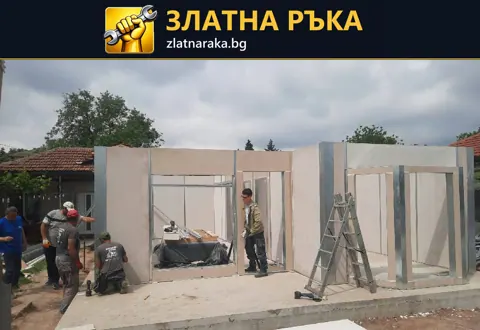 Работници монтират светли стенни панели с оформени врати и прозоречни отвори върху бетонна площадка