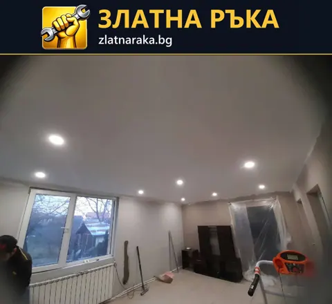 Гладък бял таван с множество малки вградени лампи над стая с прозорец и защитени мебели