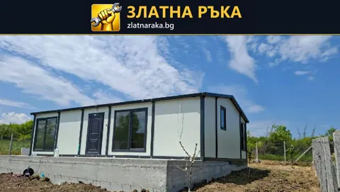 Продълговата светла сграда с тъмни рамки, големи прозорци и недовършен цокъл.
